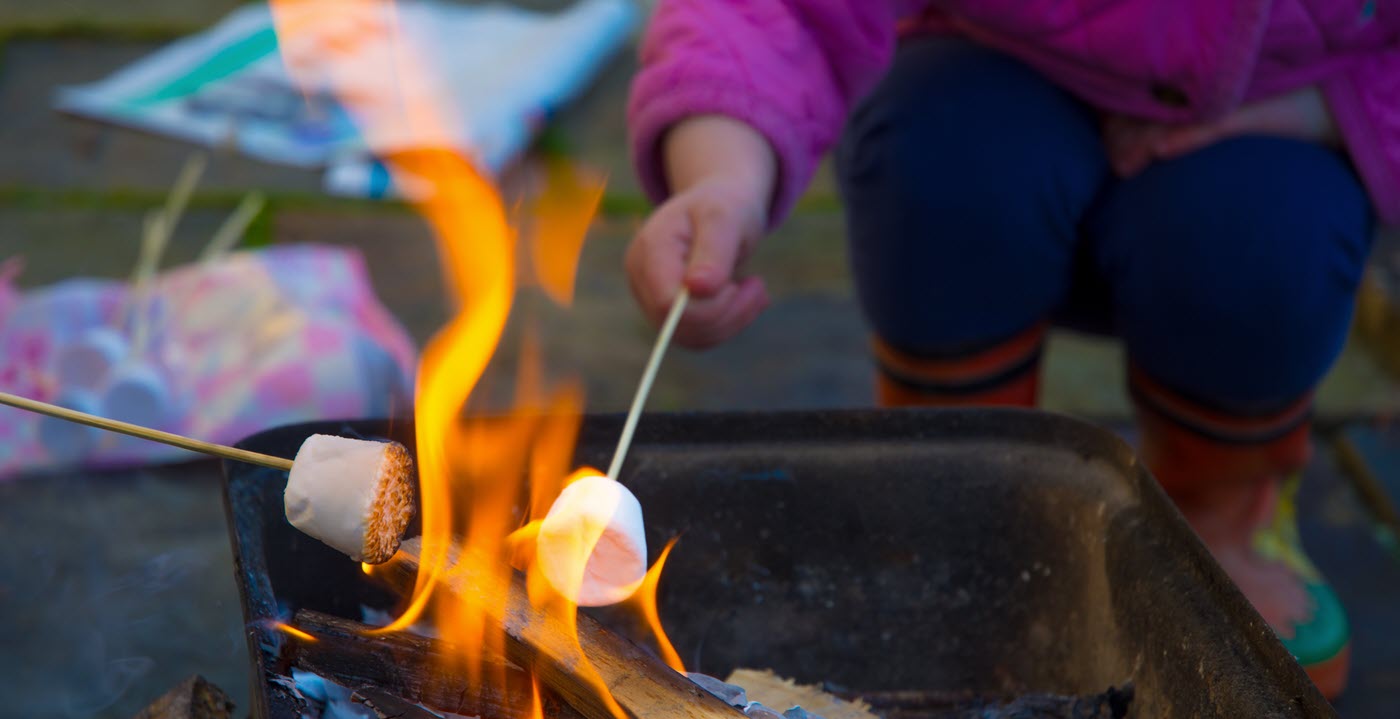 Closeup of child in pick puffy jacket and patterned boots roasting a marshmallow on a stick over a campfire.